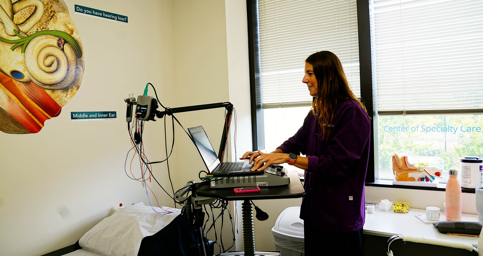 The image shows a woman sitting at a desk with various medical equipment around her, including a laptop, a microphone, and a monitor displaying health-related information.