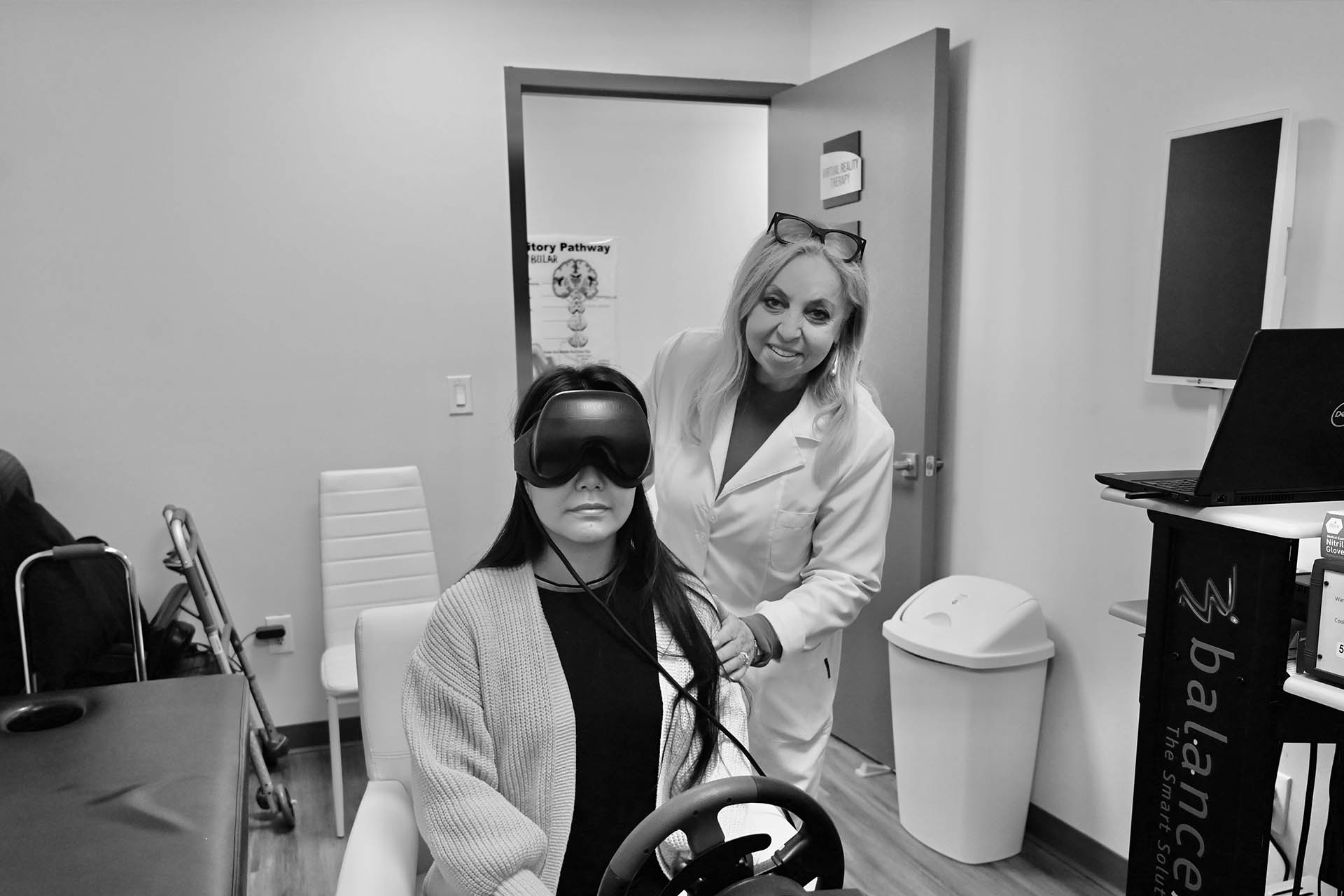A woman standing next to a person sitting in a wheelchair with virtual reality equipment on their head, both are indoors.