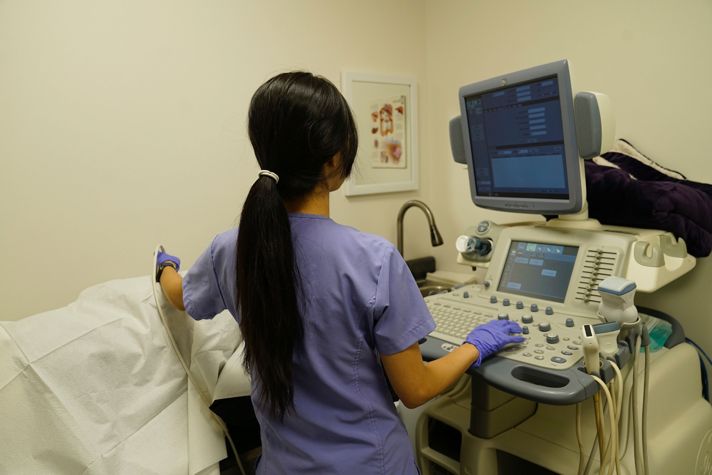 A woman in scrubs stands at an electronic medical device, possibly monitoring patient data.