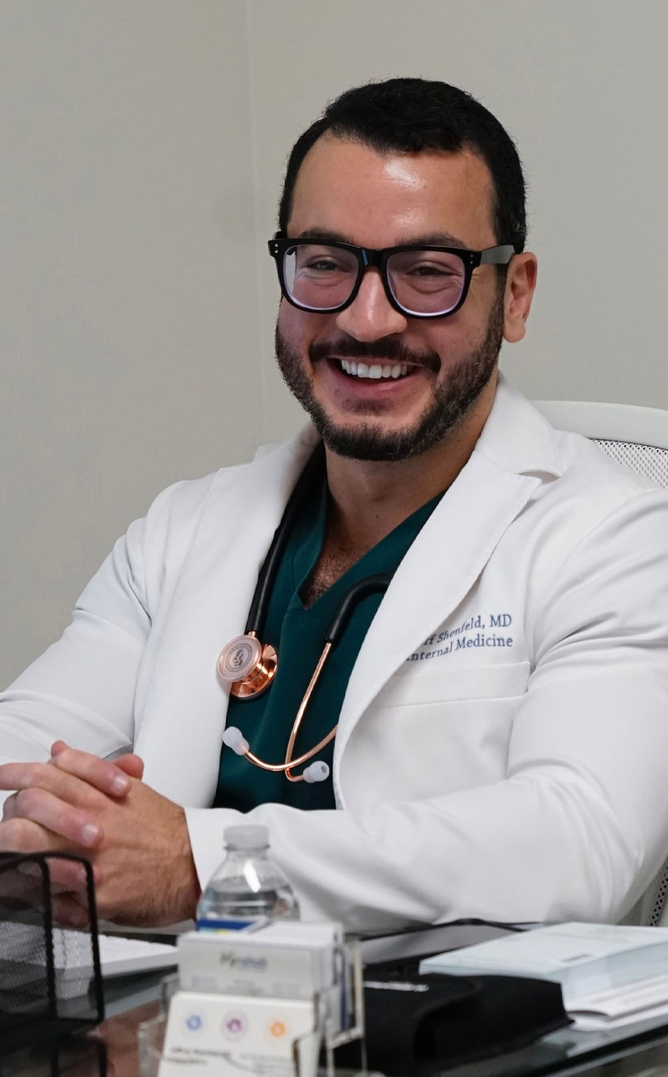 The image shows a man wearing glasses and a white lab coat with a stethoscope around his neck, smiling at the camera while seated at a desk with various items on it.