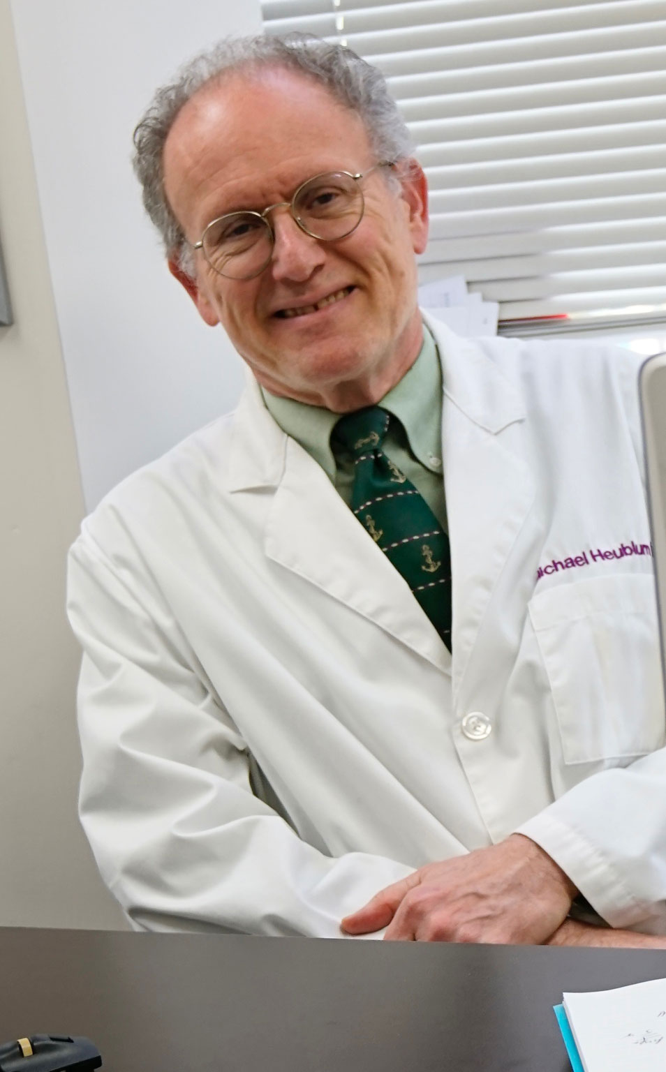 The image features a man wearing glasses, a white lab coat, and a name tag, sitting at a desk with papers, smiling and looking directly at the camera.
