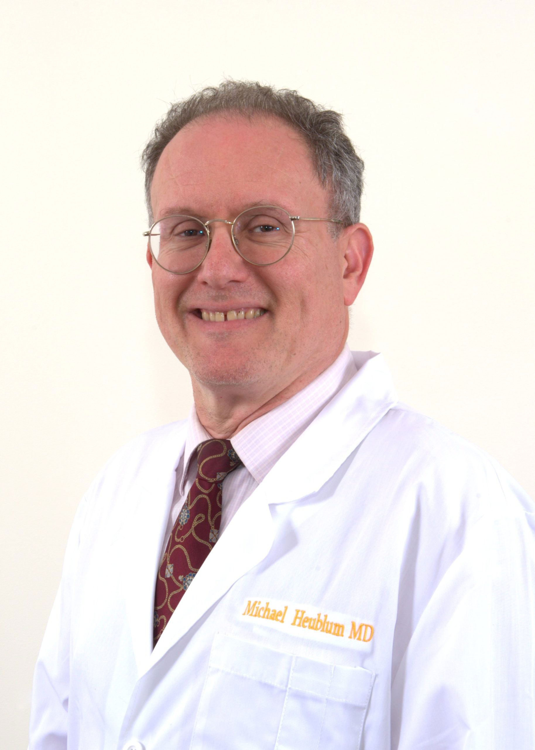 The image shows a man wearing glasses, smiling, and posing in a white lab coat with name tags, standing against a plain background.